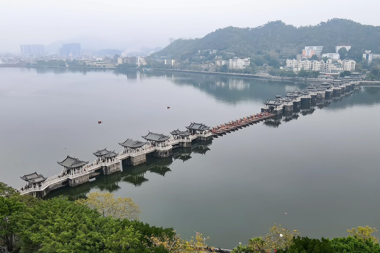 Aerial view of Guangji Bridge: the full span of stone piers and pavilions with the chained line of shuttle boats down the middle, the Han River on both sides and wooded hills beyond