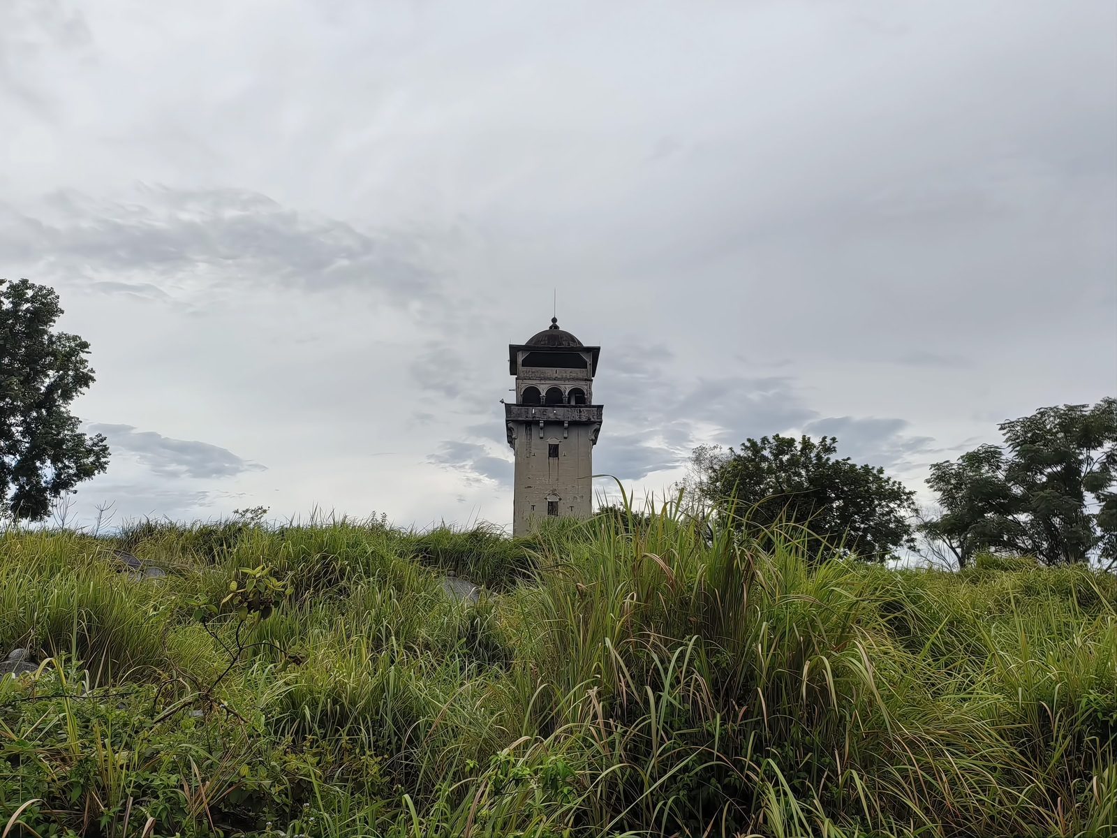 A lone five-storey concrete lamp tower standing in tall grass under a wide cloudy sky