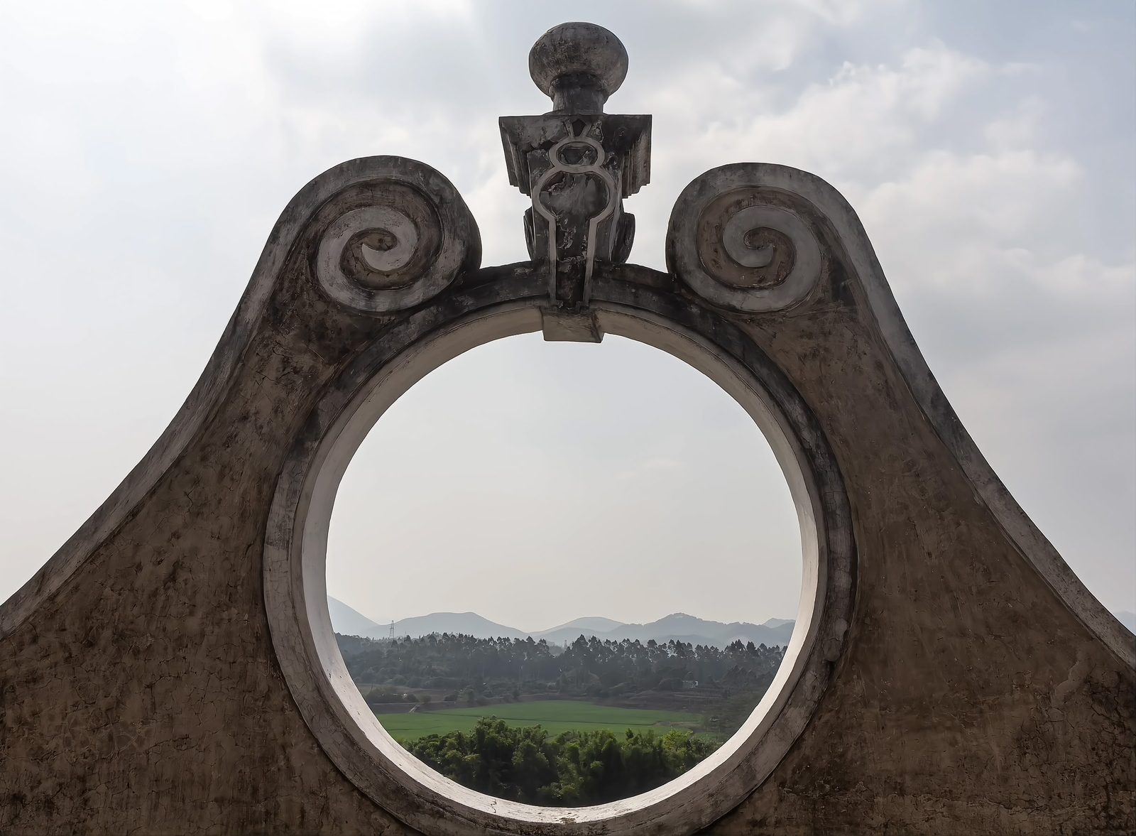A large circular window in a concrete tower framing a view of green fields and distant hills under a pale sky