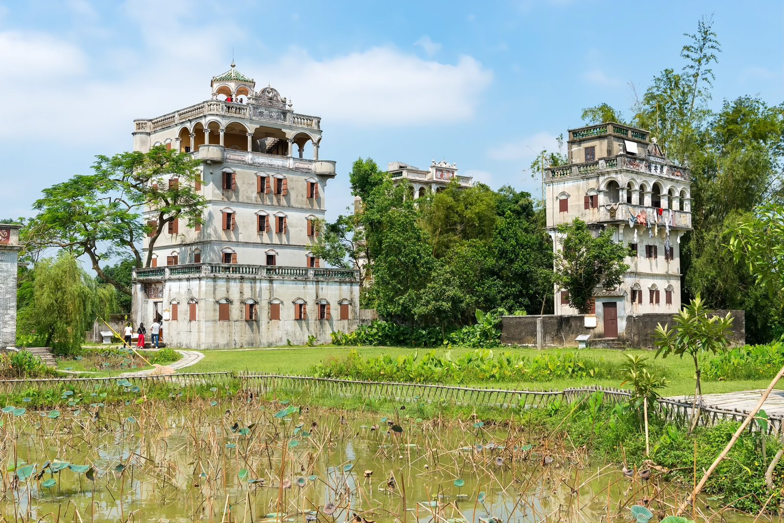 Two weathered diaolou towers standing beside a lotus pond in green countryside, with visitors walking a path between them