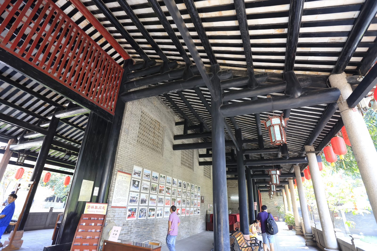 A long corridor of black timber columns and beams at Nanhai Temple, red lanterns hanging, display boards and steles along the wall, visitors resting on benches