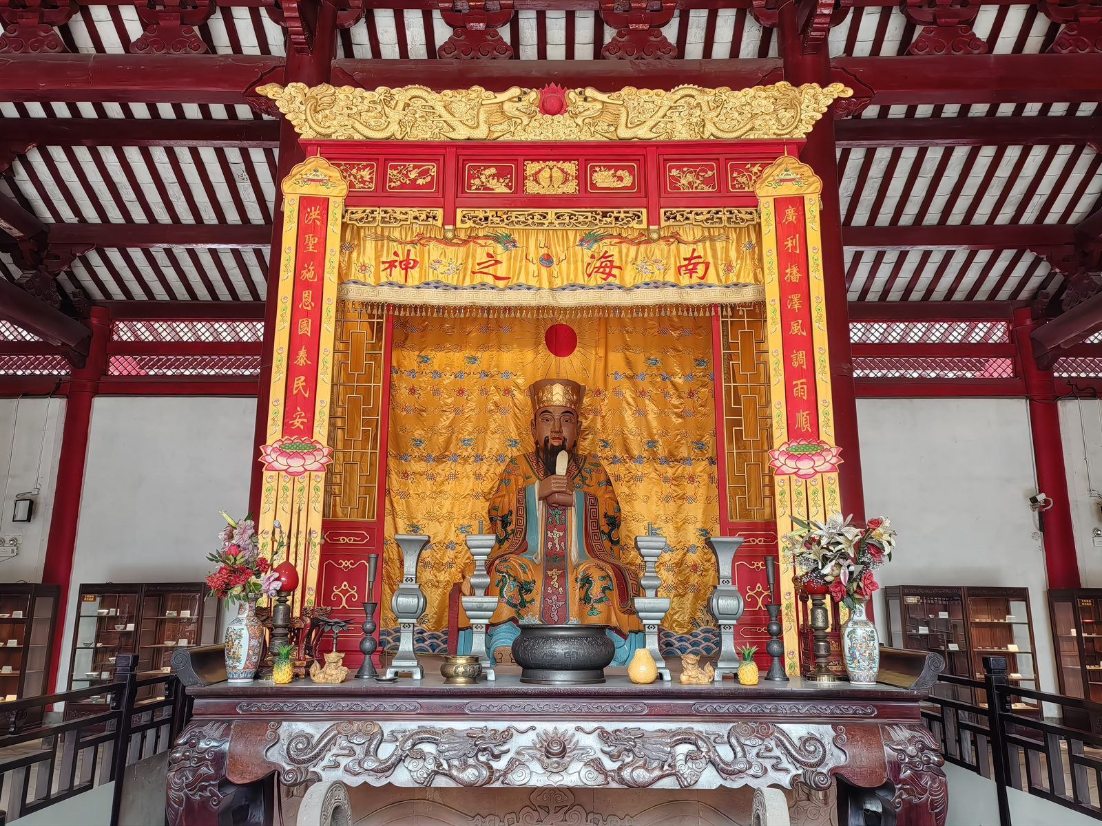 The statue of the South Sea God inside the great hall of Nanhai Temple: a bearded figure in gold robes and a gold crown seated on a throne under a yellow banner reading Nanhai zhi shen, god of the south sea, with incense burners and flower vases on the altar table in front