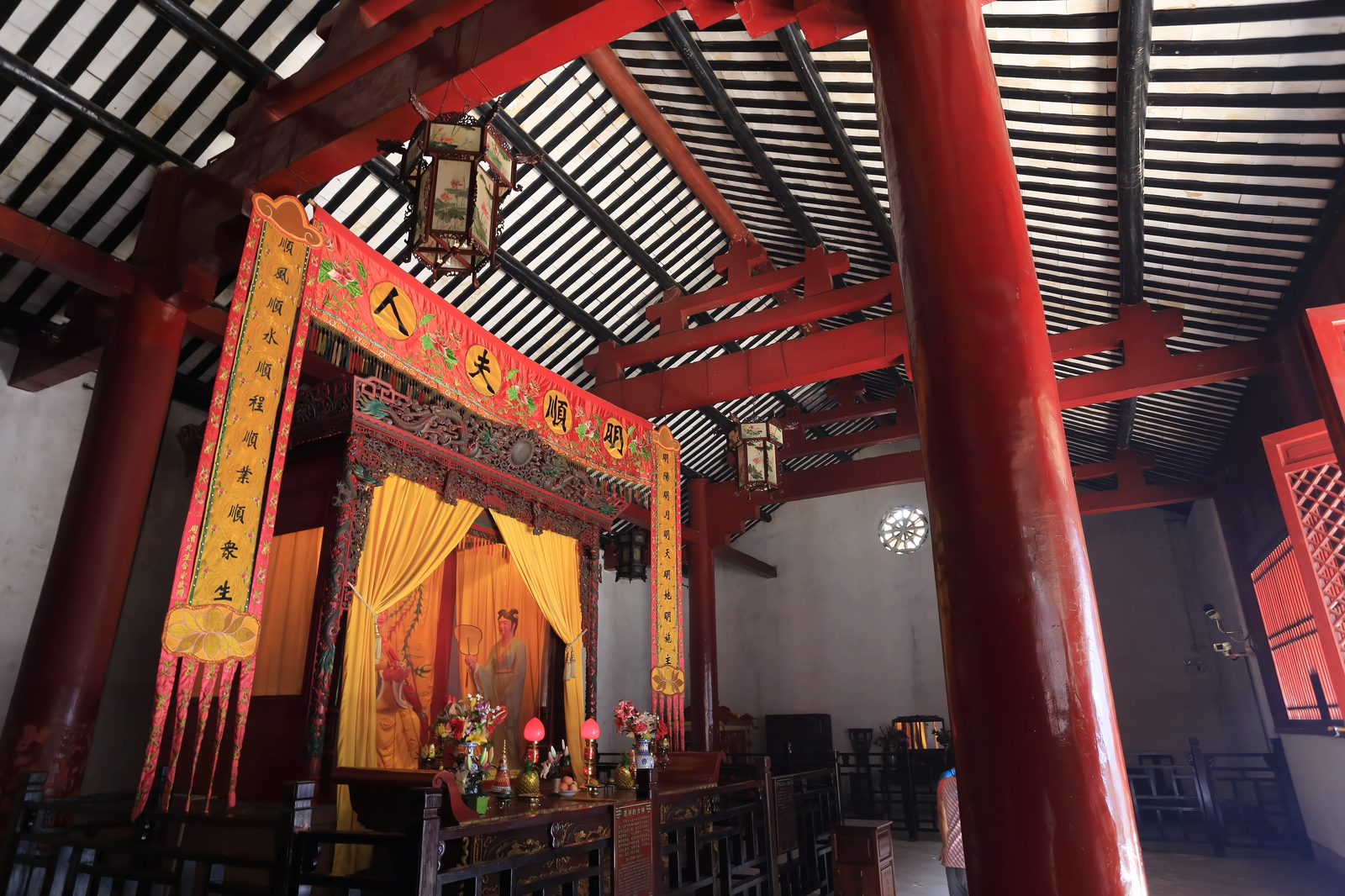 The interior of the Palace of Bright Spirits at Nanhai Temple: red columns and black timber roof, a canopied bedchamber altar with yellow curtains, and a banner reading Ming Shun Furen above the female deity