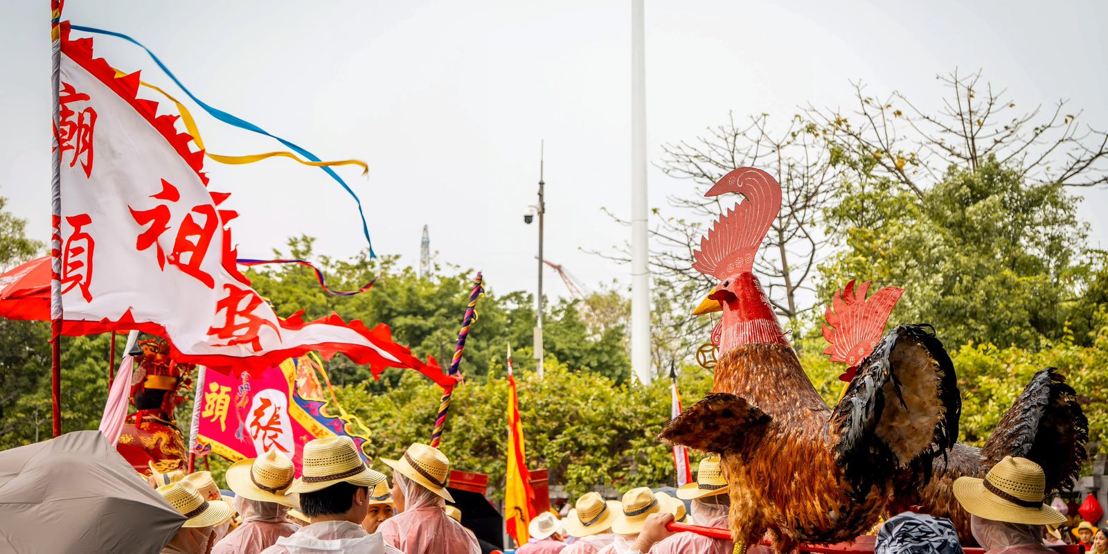Giant feathered rooster figures carried through the Boluo birthday procession at Nanhai Temple, villagers in rain capes and straw hats beneath, banners and flags above