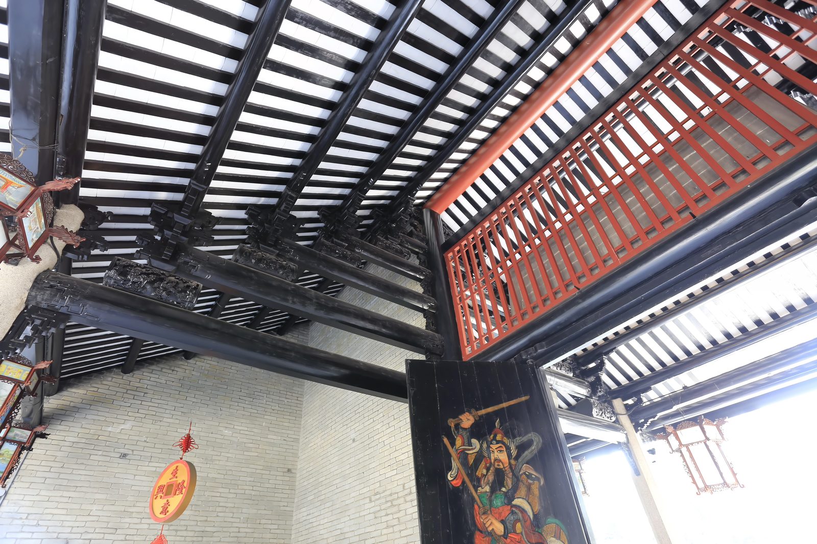 Looking up into the black timber roof structure of a gate at Nanhai Temple: heavy lacquered beams, a red lattice panel, and a painted door god with sword on the dark panel below
