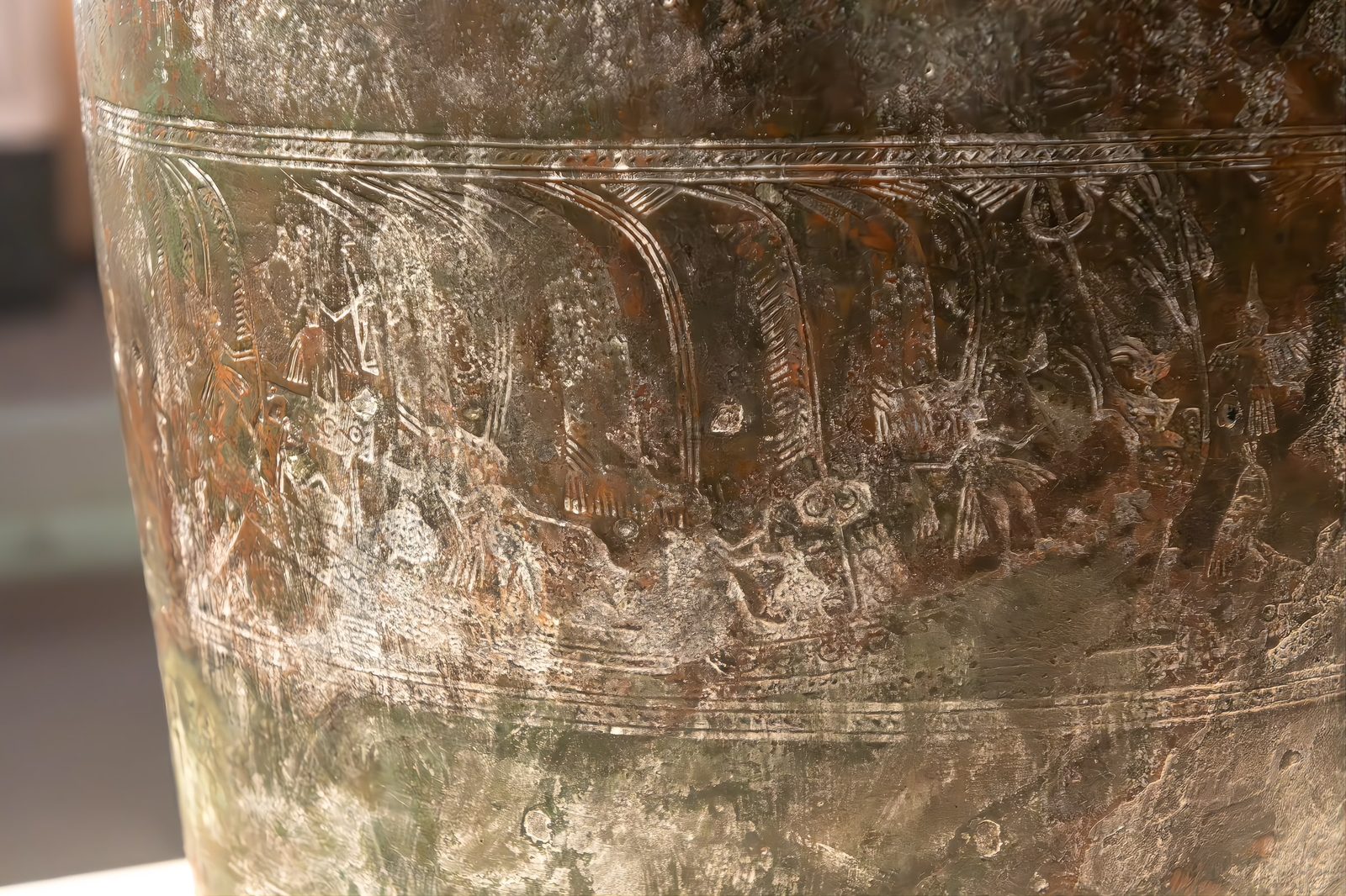 A close view of the bronze bucket's decorated upper wall, showing incised boat patterns with figures rowing, worn patina across the bronze surface
