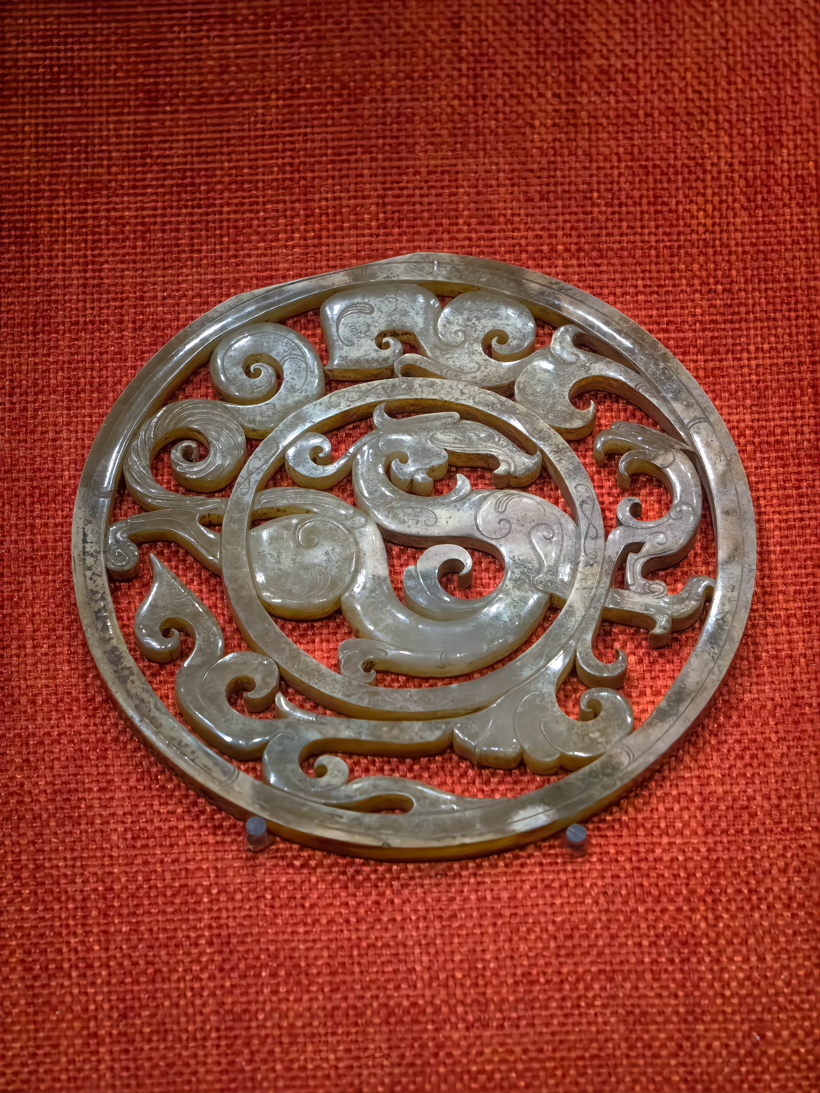 A grey-green jade openwork disc carved with an intertwined dragon and phoenix pattern, displayed against a dark red background