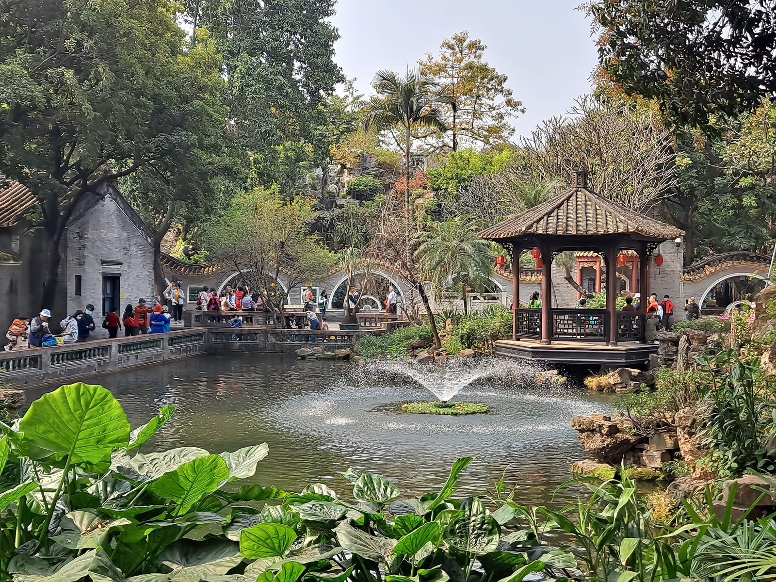 A hexagonal pavilion standing in a pond at Qinghui Garden, reached by a stone bridge, with trees and traditional buildings around the water