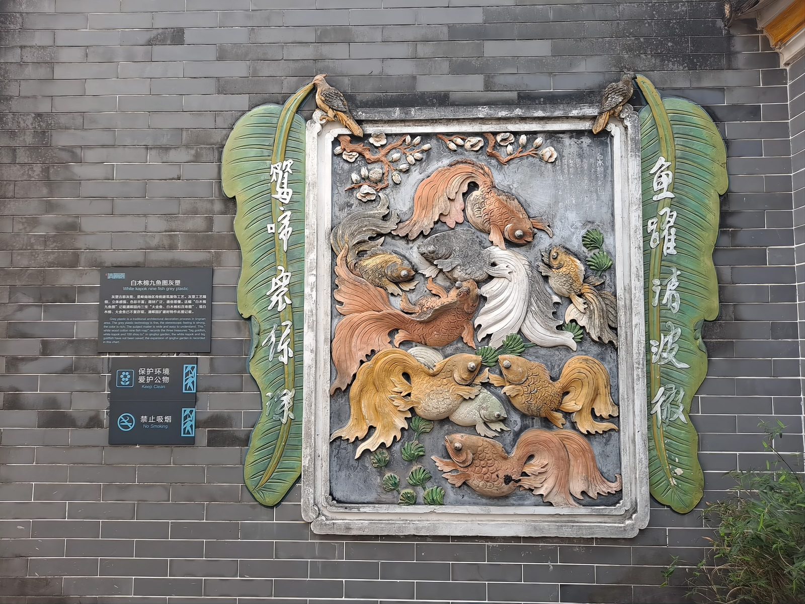 A lime-plaster relief on a grey brick wall: nine fish swimming among weeds and waves, modelled in high relief and painted in muted greens and ochres