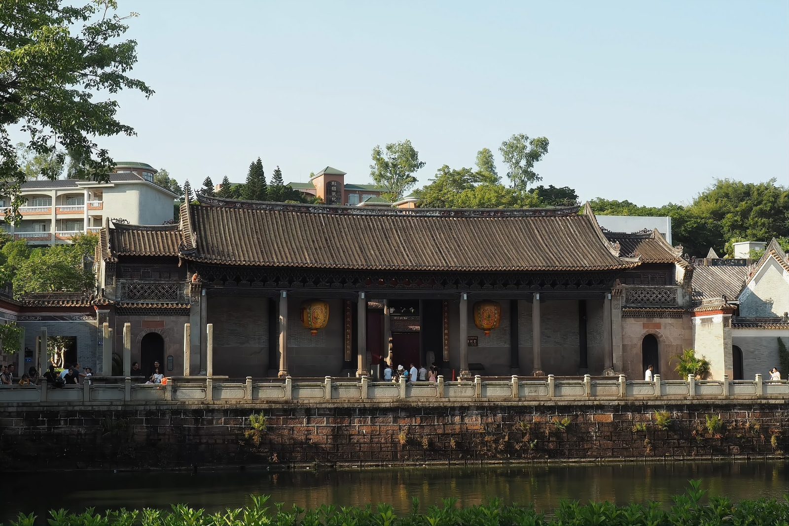 The Liugeng Hall, the great ancestral hall of the He clan, seen across a still pond in warm late light, its long tiled roof mirrored in the water