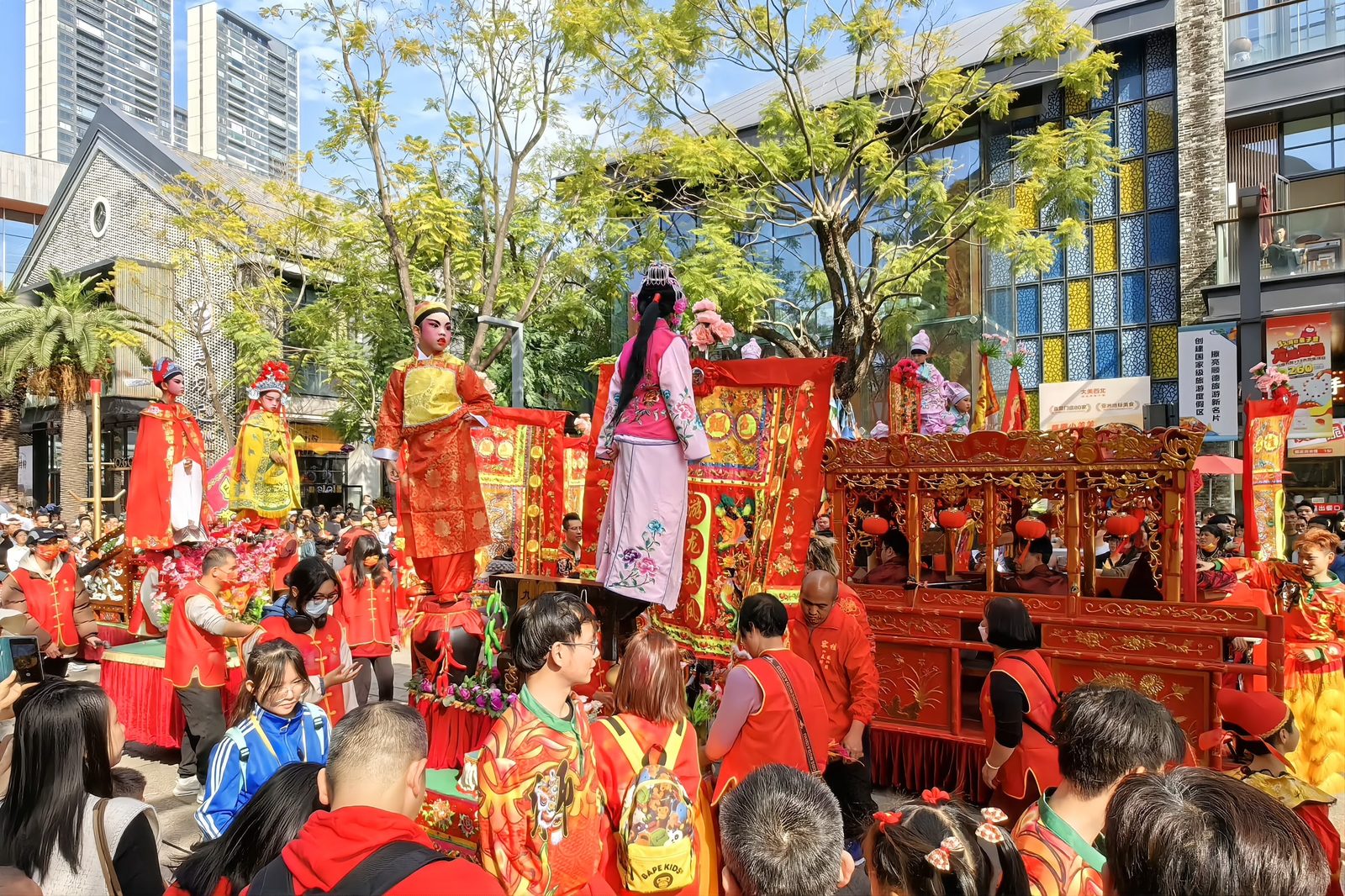 A Lingnan floating-colours parade: children in opera costume standing and seated on tall decorated cabinets, carried through a crowd between red banners