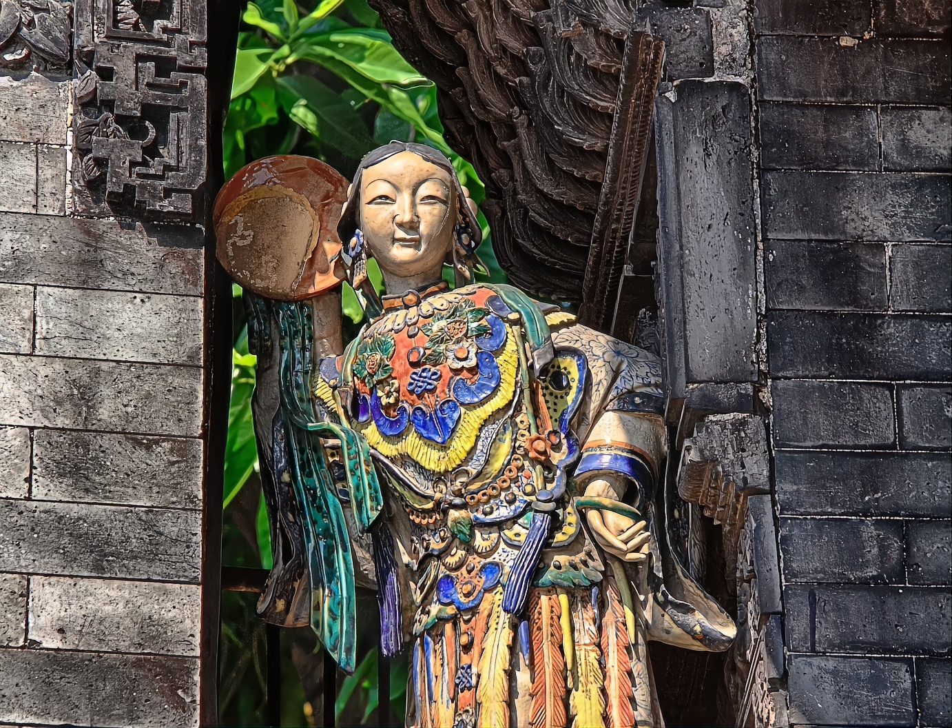 A single large ceramic figure of an opera character holding a plate, standing under dark carved eaves against grey brick
