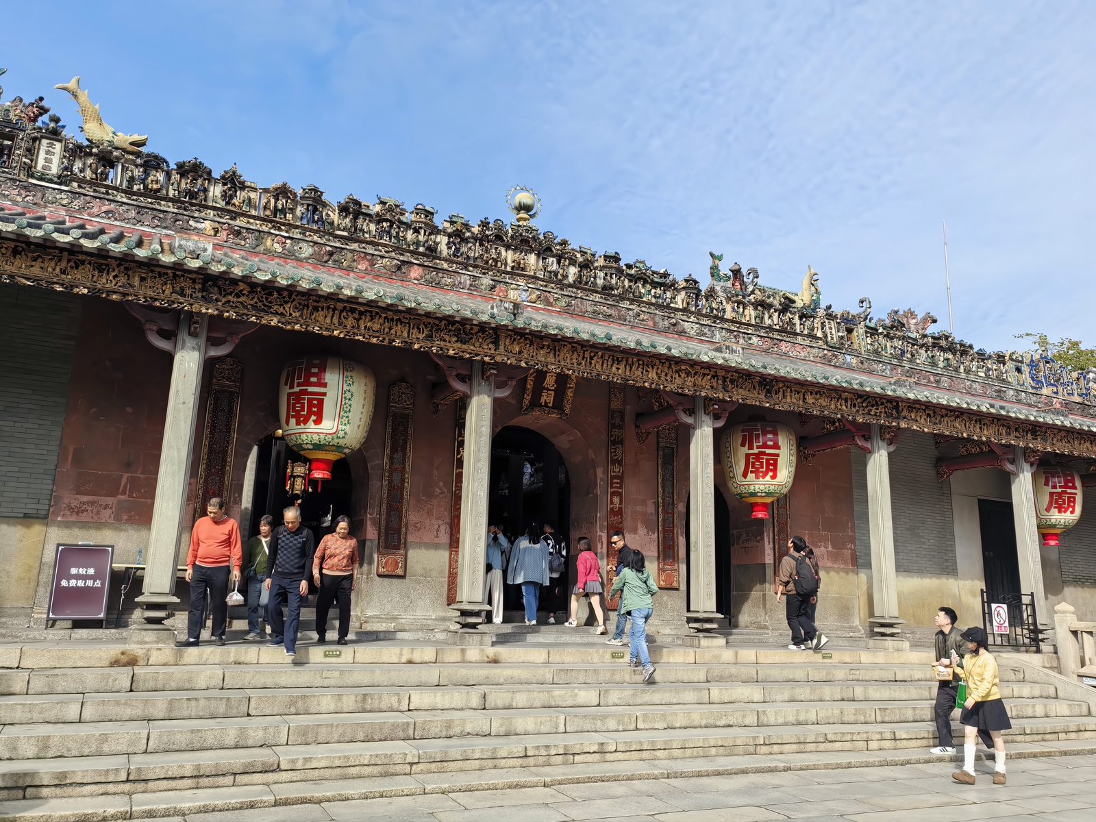 The Three Gates of the Foshan Ancestral Temple: a long grey-brick facade under a tiled roofline crowded with hundreds of small ceramic figurines, red-and-gold lanterns reading Zumiao hanging between the columns, visitors on the stone steps