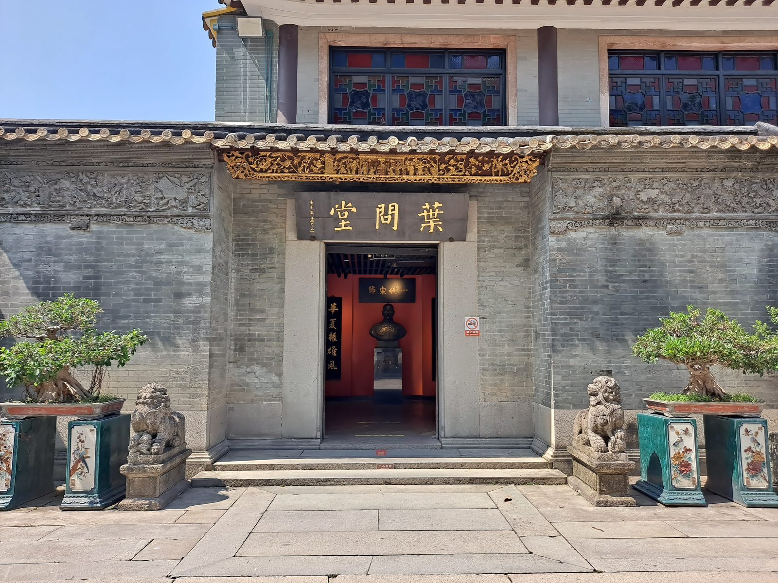 The entrance of the Ip Man Hall at the Foshan Ancestral Temple: a grey-brick gatehouse with a gold name board reading Ye Wen Tang, two stone lions and potted penjing flanking the doorway, and a bust of Ip Man visible inside