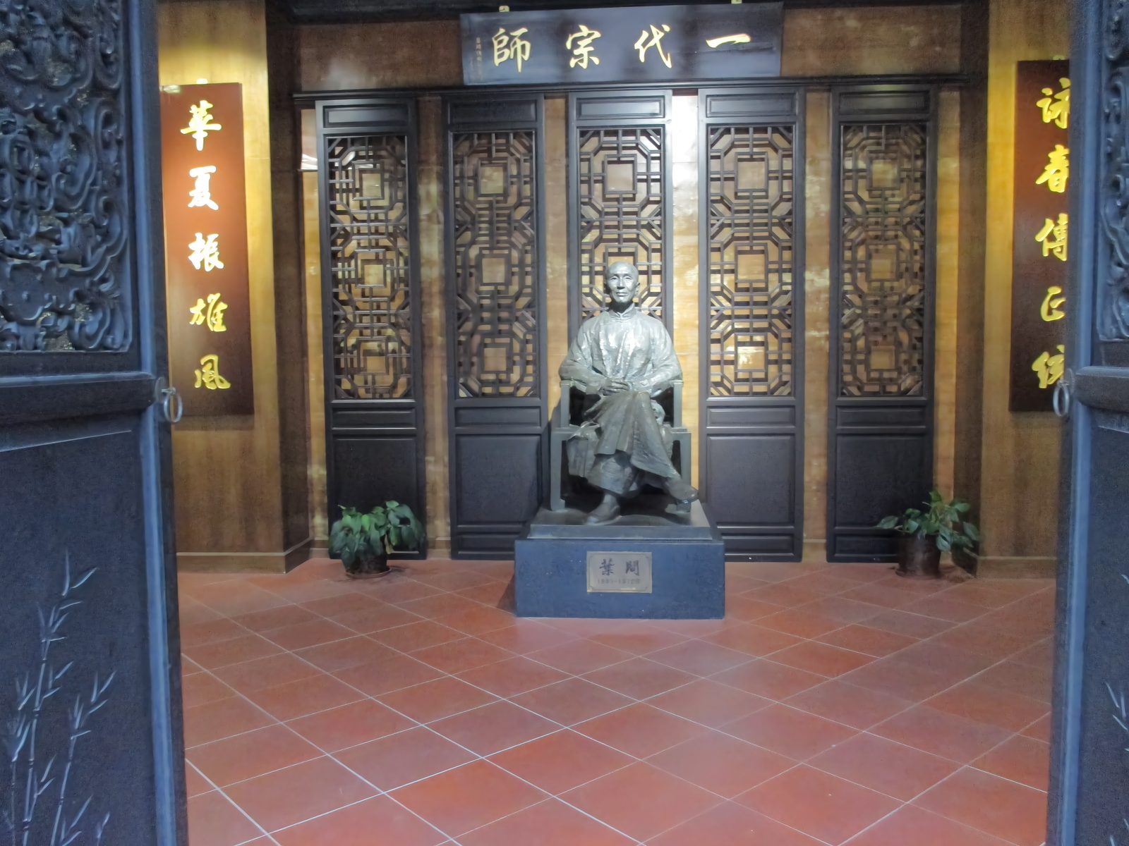A seated bronze statue of Ip Man inside his memorial hall at Luo Village, Foshan, under a plaque reading Yi Dai Zong Shi, patriarch of a generation, with lattice screens and a red tiled floor