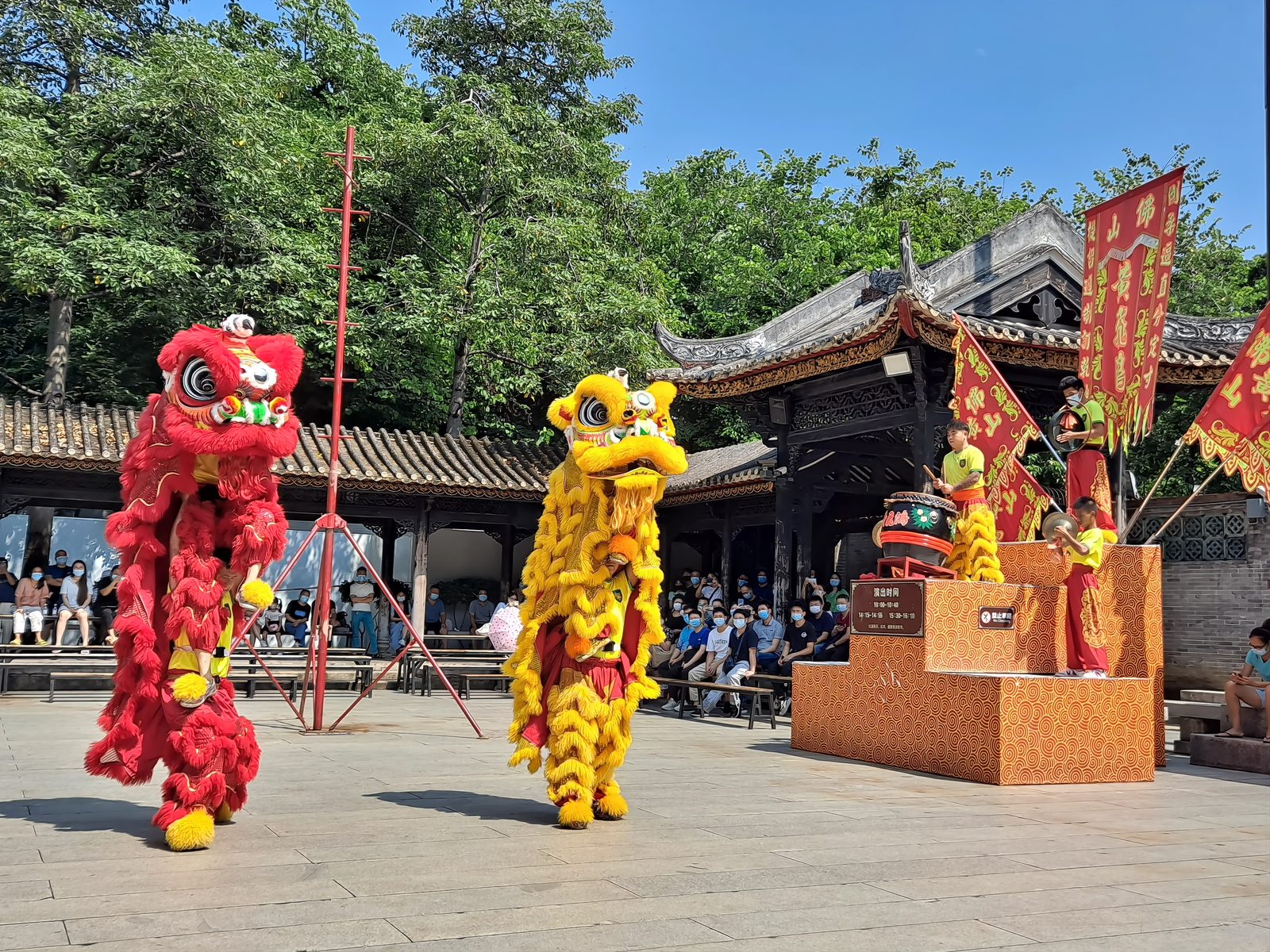 A red lion and a yellow lion performing in front of the Wong Fei-hung Memorial Hall at the Foshan Ancestral Temple, drummers on a raised platform behind, a crowd watching