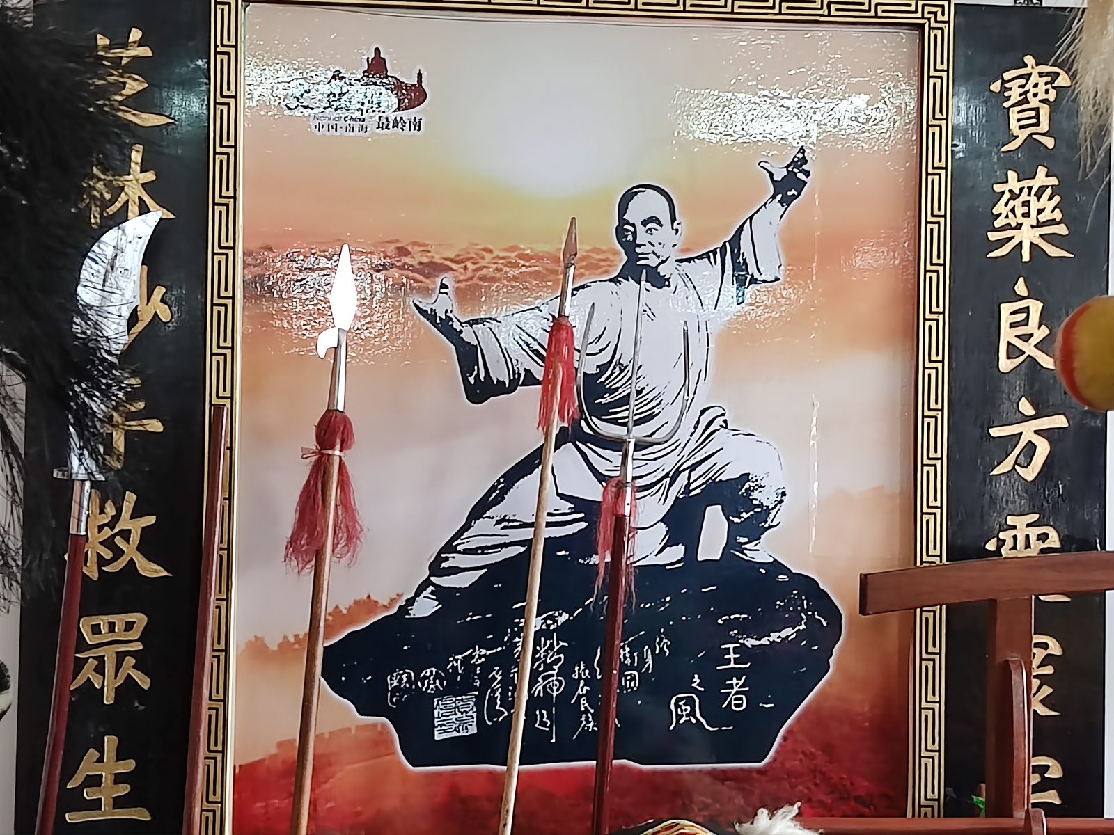 A framed portrait of Wong Fei-hung in a pale jacket striking a spear pose on a red wall under a gold name board reading Bao Chi Lam, weapons racked beside it and a lion head in the foreground