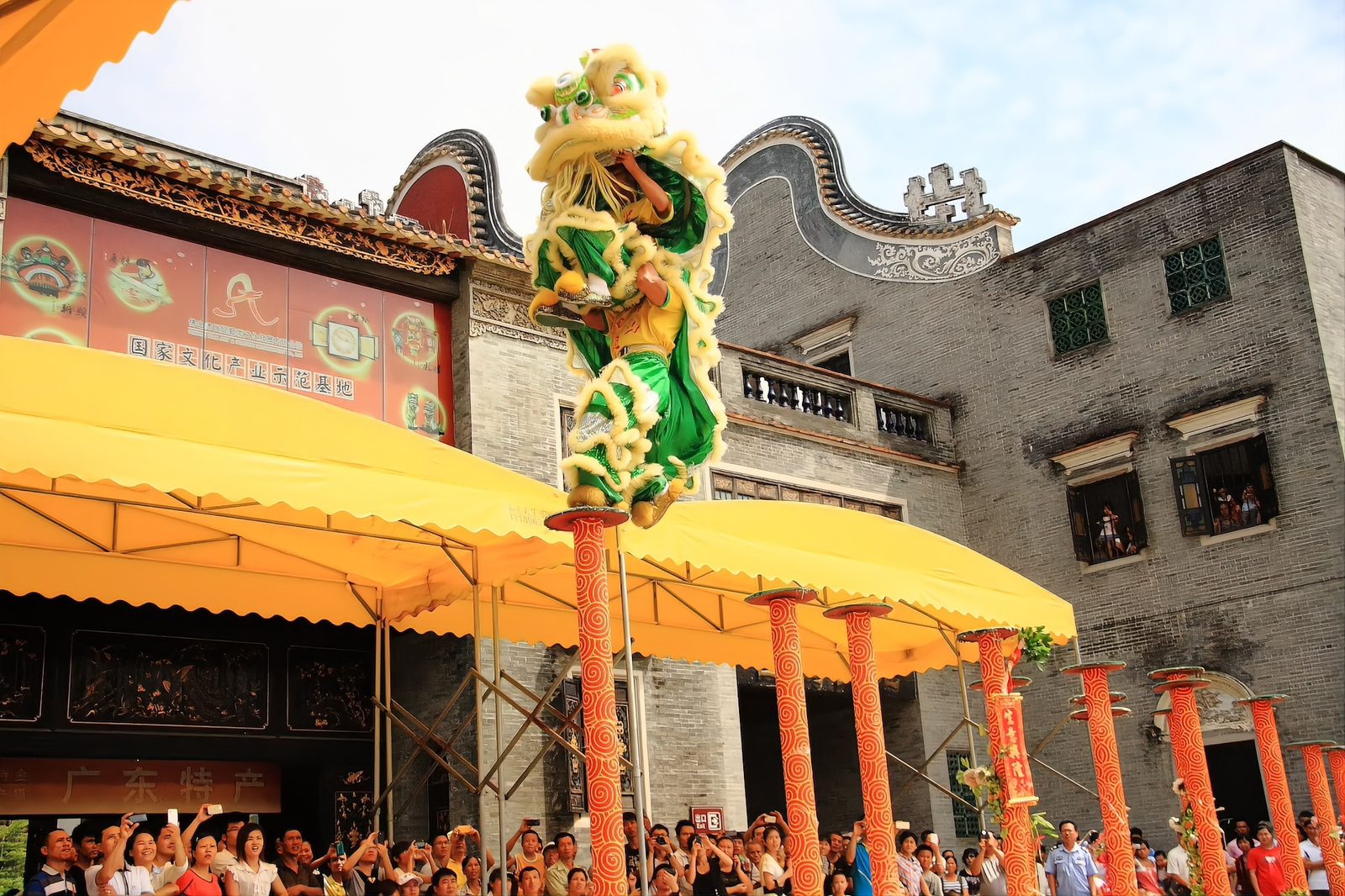 A green and gold lion climbing a row of painted poles in front of a Lingnan building, a crowd below with phones raised, the lion balanced on a pole at the top of its climb