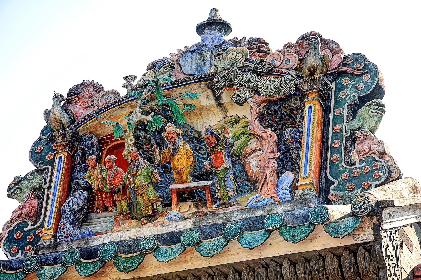 Close view of the ceramic roof frieze: a group of glazed opera figures in yellow, green and red robes around a table, with trees and birds, under a curved ridge of teal glazed tiles
