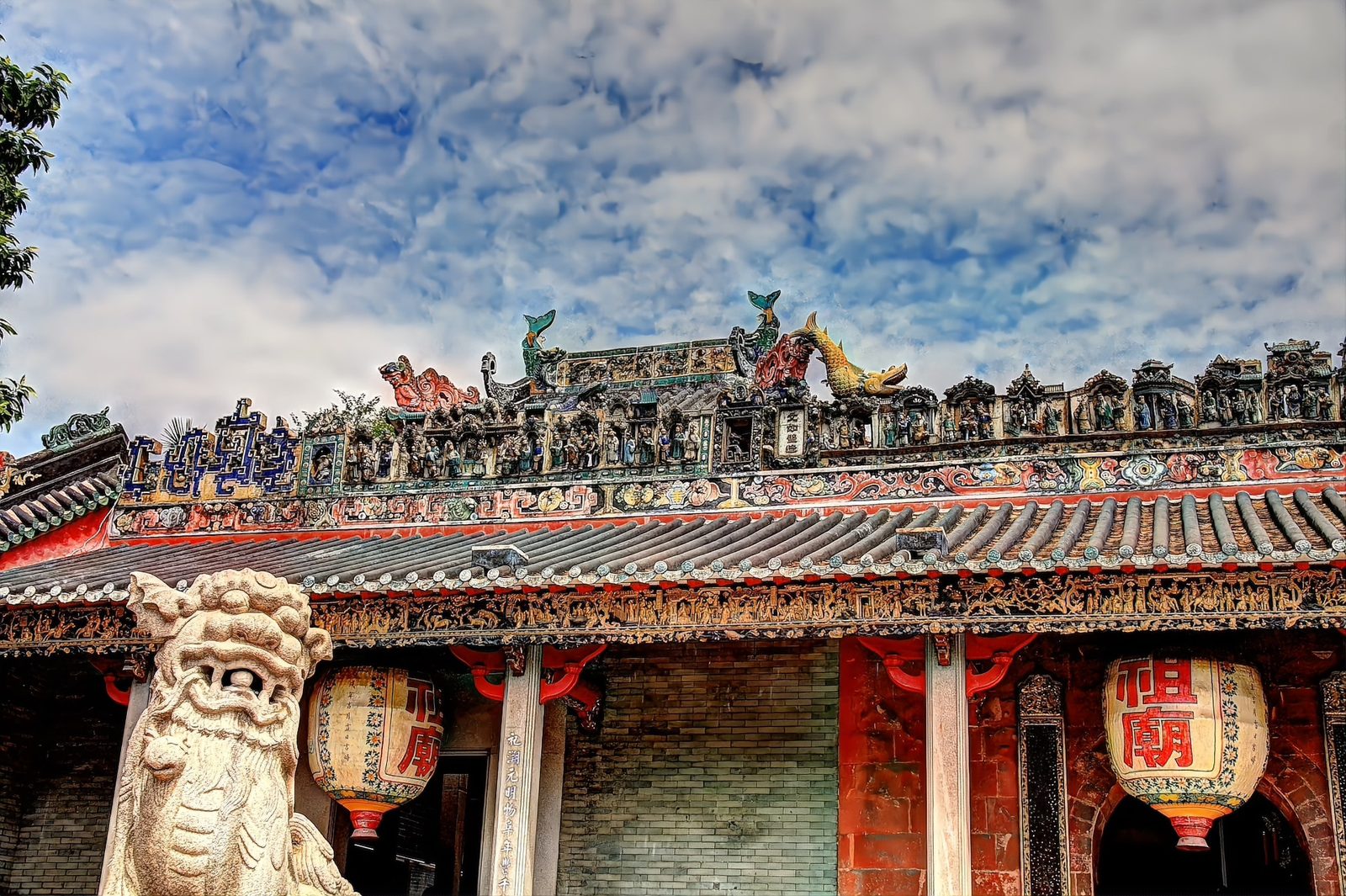 Wide view across the Ancestral Temple roofline: a stone lion in the foreground, rows of ceramic figures along the roof ridge, and red-and-gold Zumiao lanterns under the eaves
