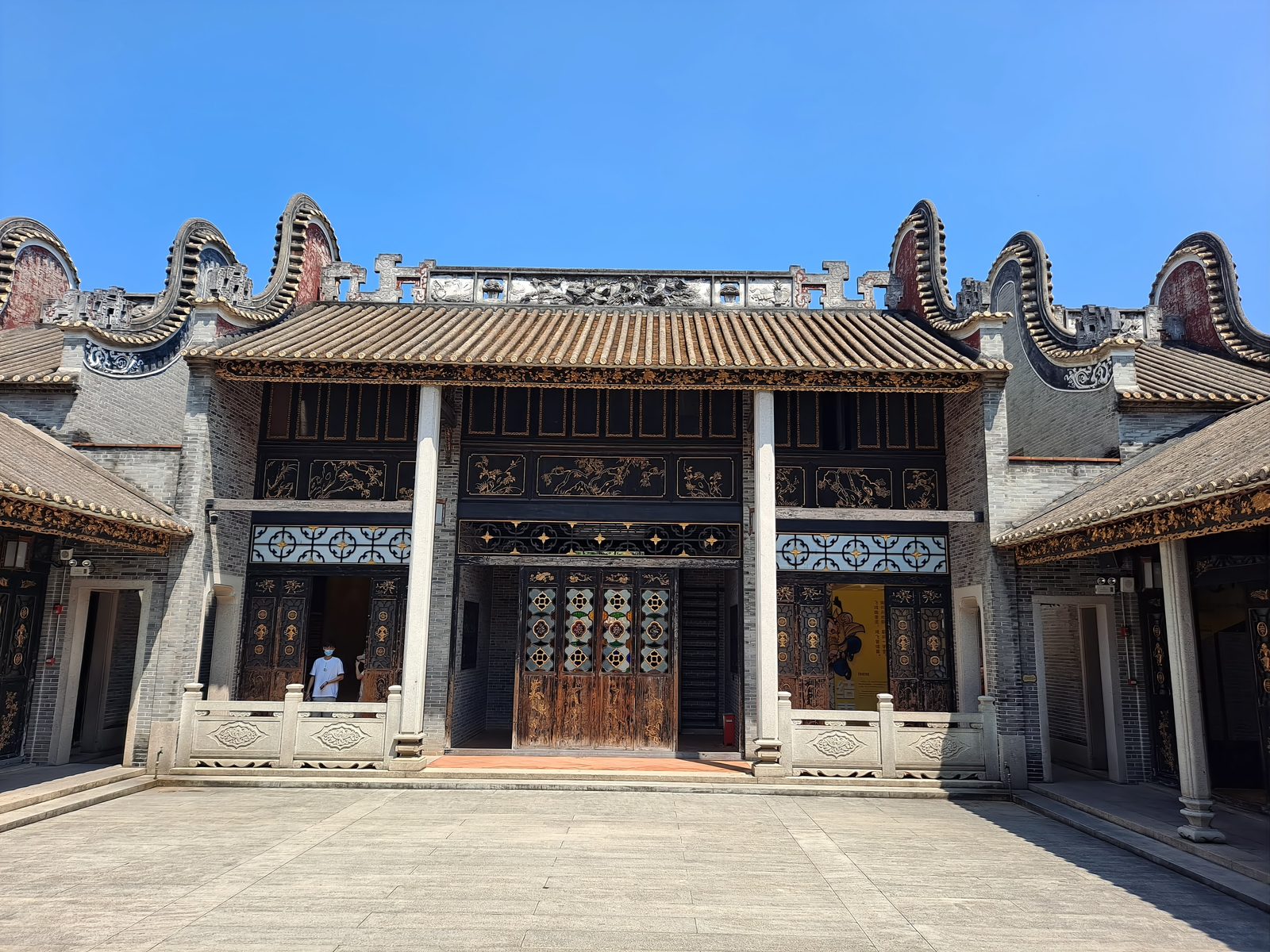 The Wong Fei-hung Memorial Hall at the Foshan Ancestral Temple: a two-storey Lingnan courtyard building with a wok-ear roofline, black-timber doors and painted girders, an empty stone forecourt in front
