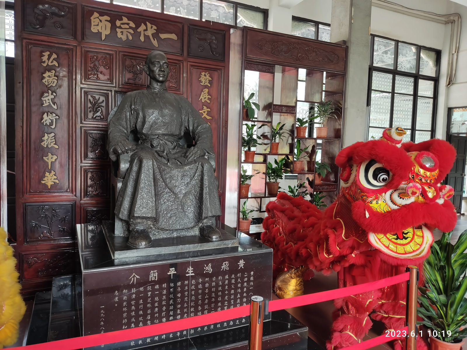 A large seated bronze statue of Wong Fei-hung at the memorial hall on Xiqiao Mountain, Foshan, under a plaque reading Yi Dai Zong Shi, with a red lion head at its side and a rack of weapons behind