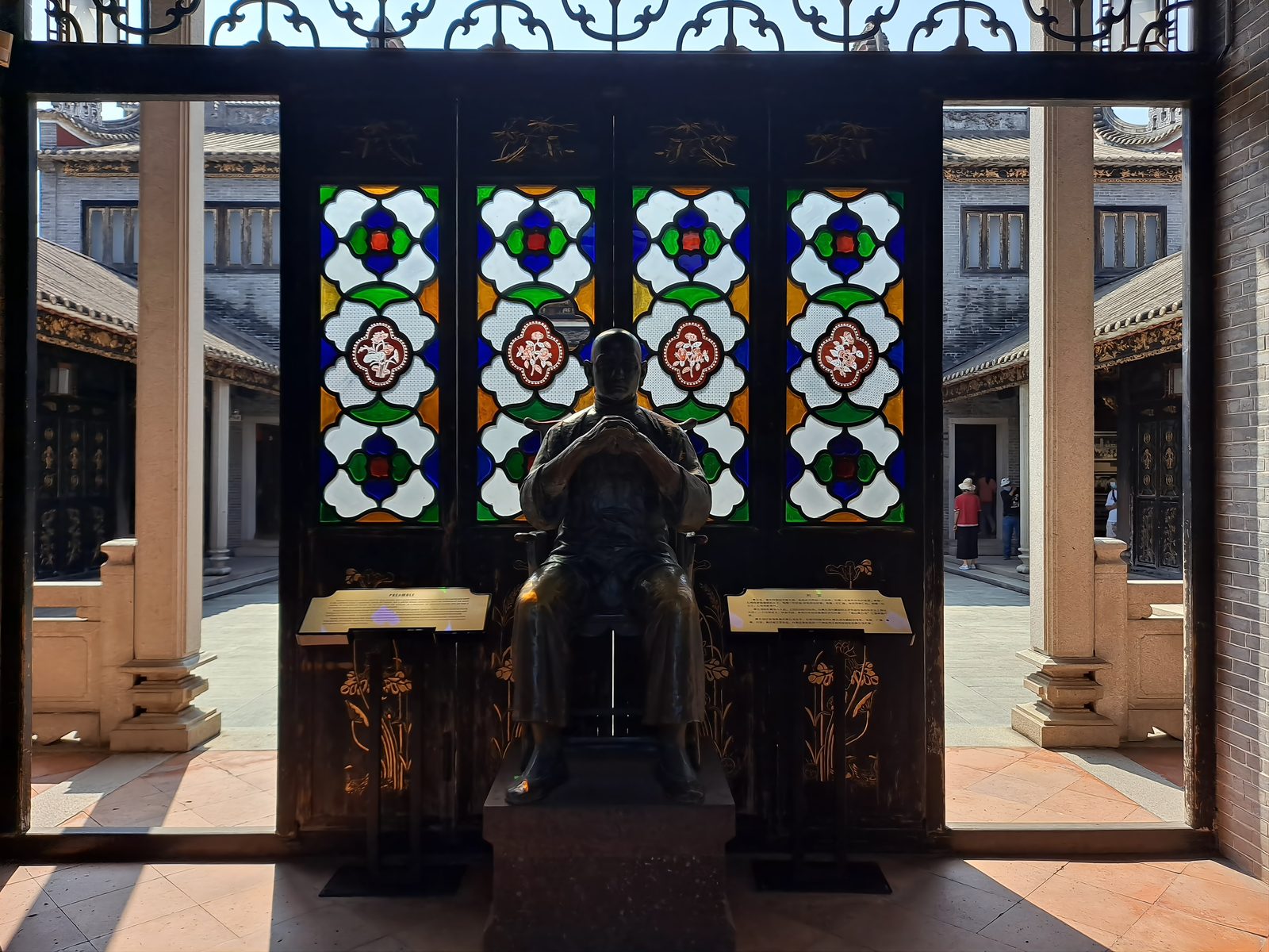 A bronze seated statue of Wong Fei-hung with hands clasped in a martial salute, standing in a doorway of stained-glass panels inside the Wong Fei-hung Memorial Hall at the Foshan Ancestral Temple